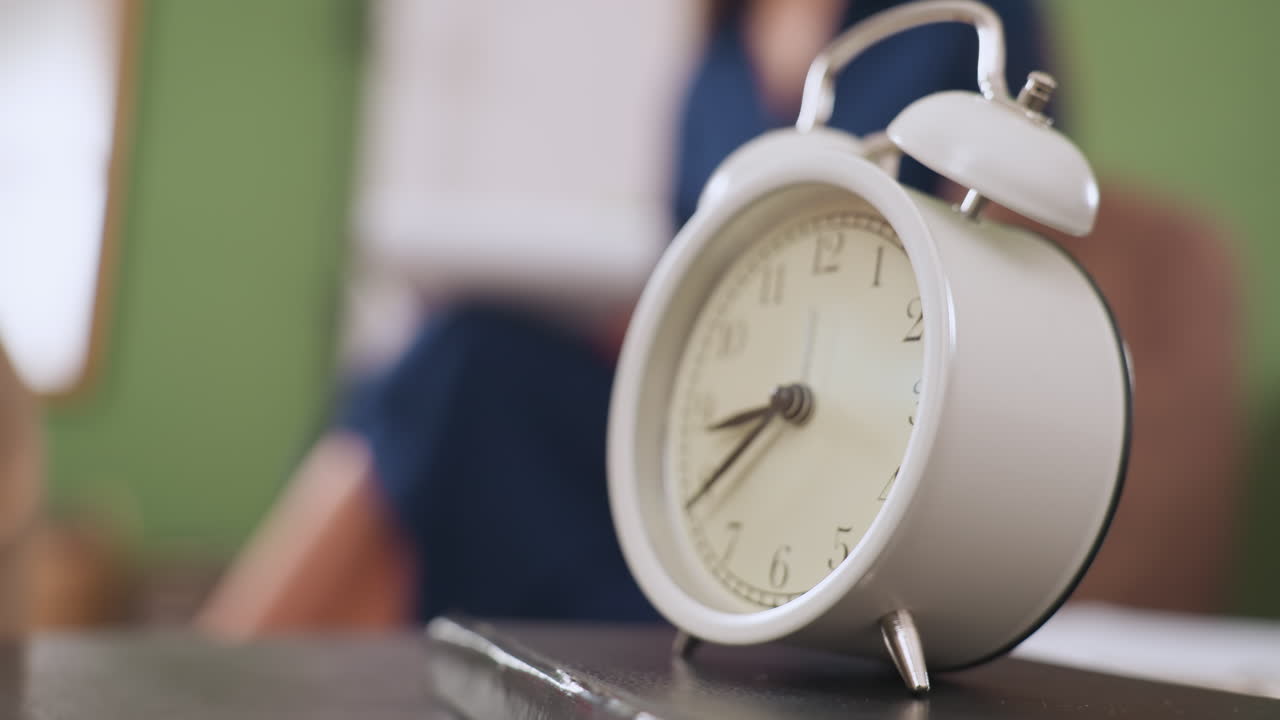 Classic white clock placed on table over notebook in foreground, with blurred view of specialist seated on sofa holding pen and writing note in calm contemplative room setting