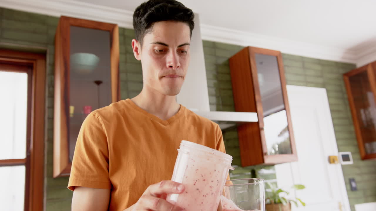 hombre biracial enfocado vertiendo batido de frutas de la licuadora y bebiendo en la cocina, cámara lenta