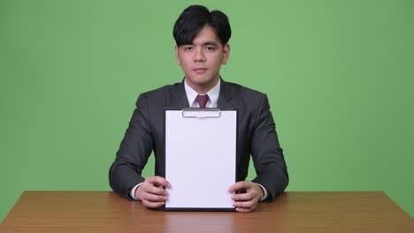 young handsome asian businessman working with clipboard against green background