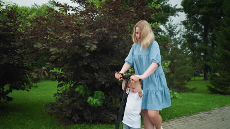 a mother and her young son share a scooter ride along a tree-lined pathway, the mother uses her leg to propel the scooter as they move forward