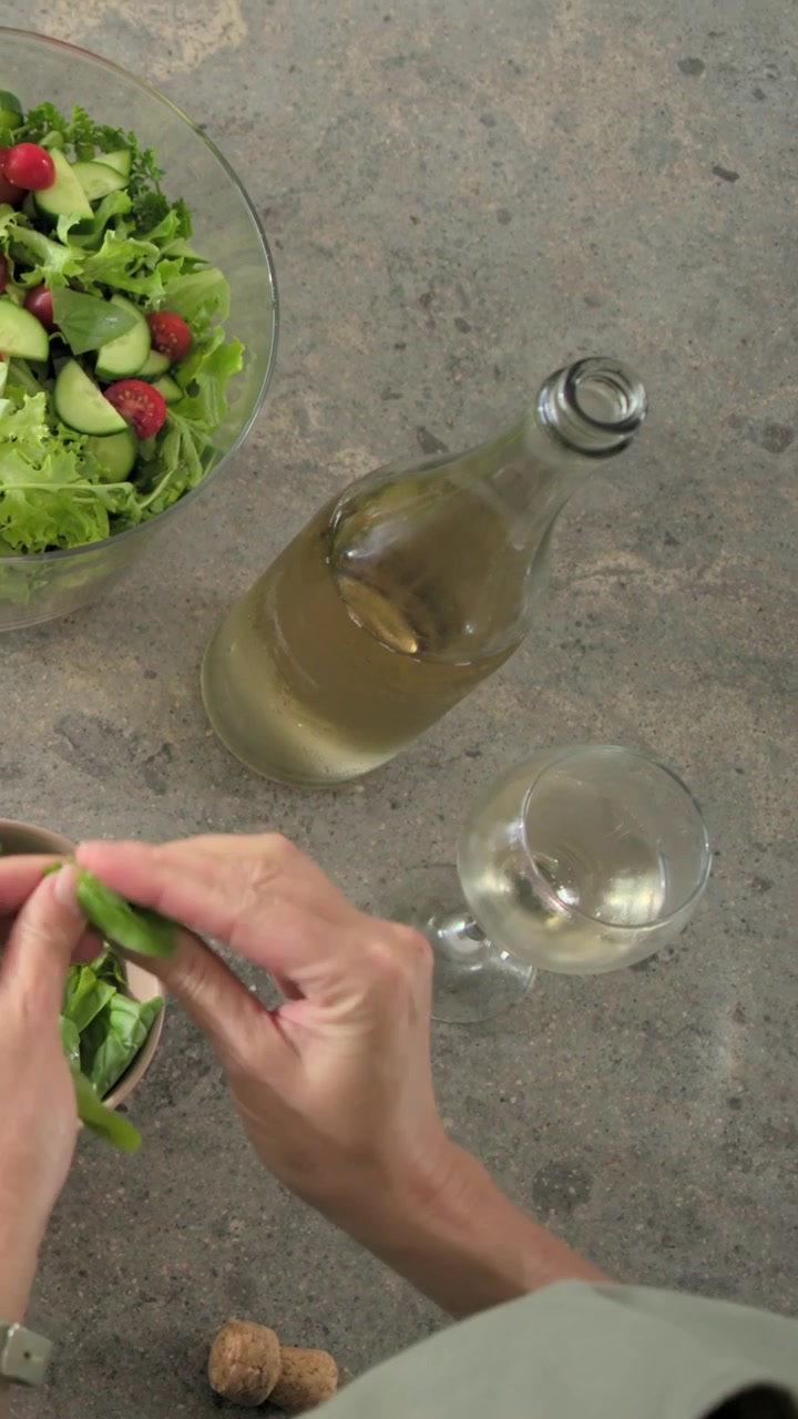 Video vertical: Mujer con top claro terminando la ensalada, arrancando albahaca, sirviendo vino en la encimera, reloj.