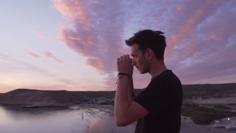 jonge man die op de berg staat en een zonnebril opzet terwijl hij naar de zee kijkt bij zonsondergang