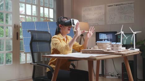 side view of smiling asian woman working with a model of small house roof solar panel wearing vr and showing touching gesture