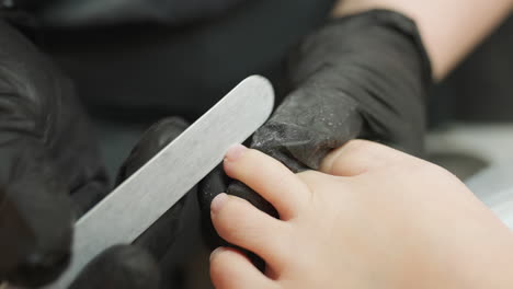 technician wearing black gloves carefully files client toenails with visible dust particles on gloves, while client leg rests on cushioned bed during detailed pedicure in clean professional setting