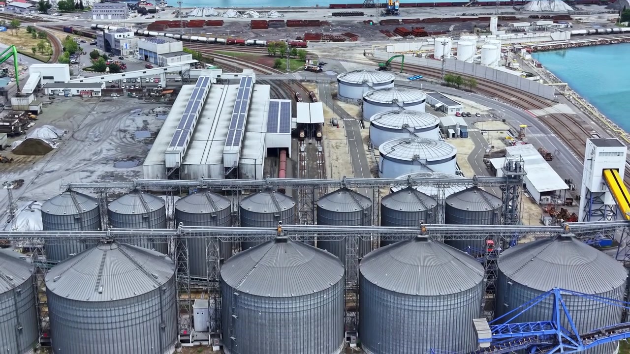 Ansicht von industriellen Lagertanks und Eisenbahnschienen in der Nähe eines Hafens in einem Küstengebiet, mit Frachtschiffen im Hintergrund, tagsüber.