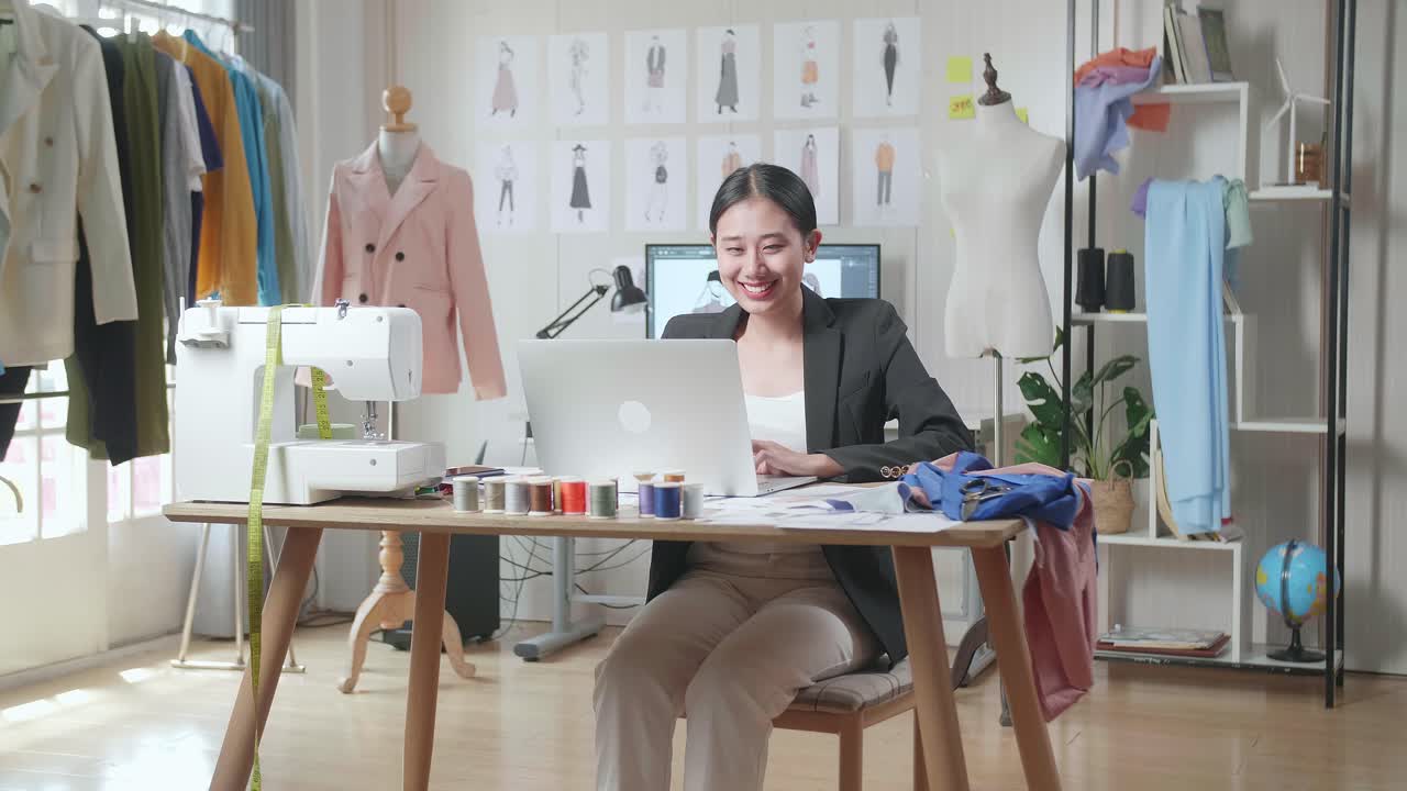 스튜디오에서 옷을 디자인하는 동안 노트북에서 일하는 비즈니스 슈트에 미소 짓는 아시아 여성 디자이너