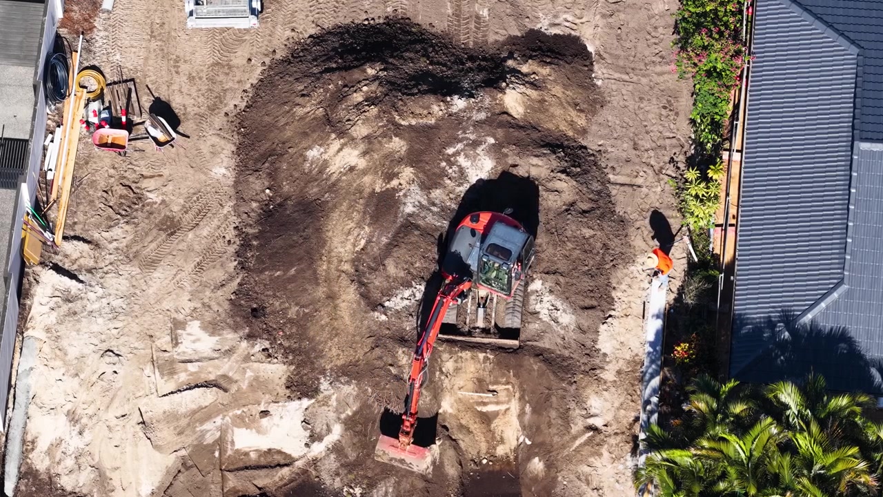 Top-down drone shot of an orange excavator moving soil in a sunny backyard construction area.