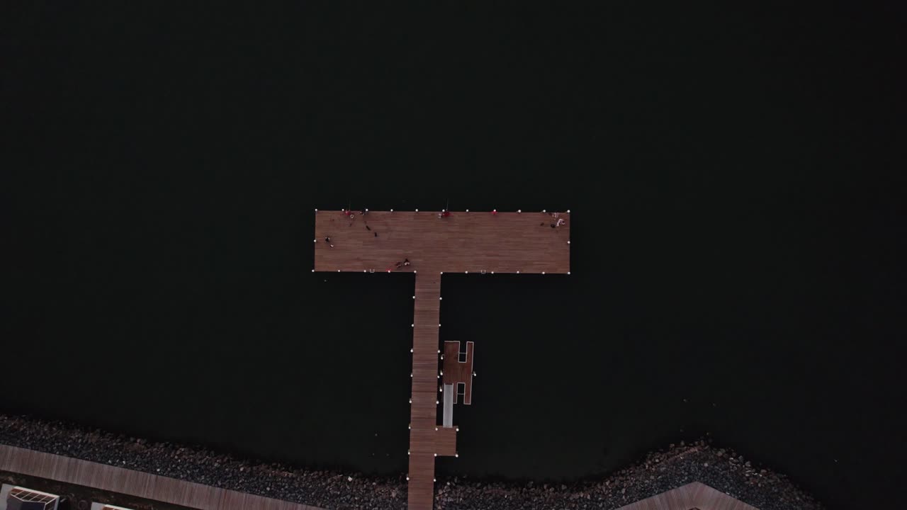 Drone top down ascend above T shaped wooden dock pier with fishermen along boardwalk with homes
