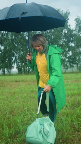 woman holds an open umbrella in one hand and bag in the other, walking in a grassy field, she drops the bag on the ground while, with a background of tall trees and a vast field