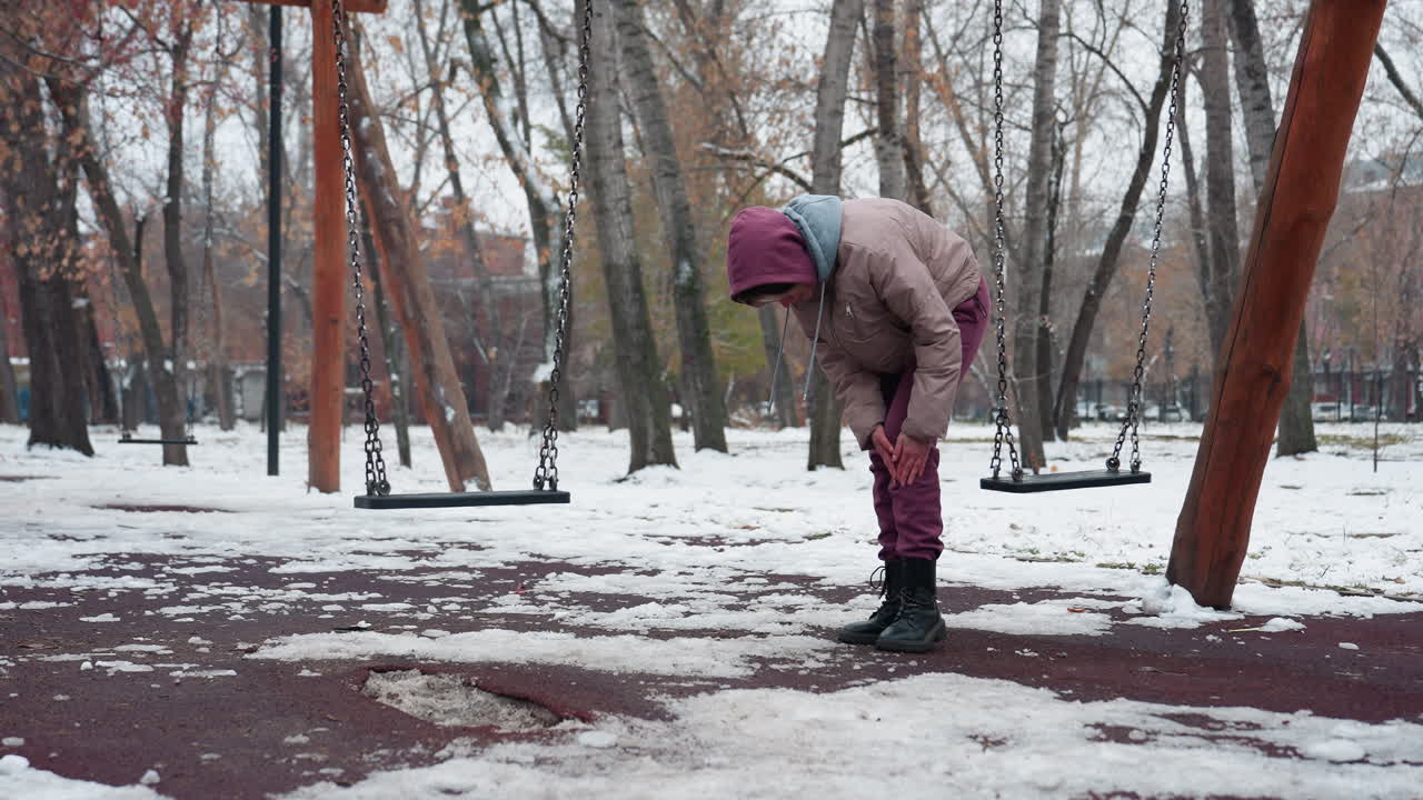 겨울 옷을 입은 여자가 무을 구부리고 을 때 무에 통증이 느지며, 벌거벗은 나무와 눈으로 인 땅의 배경으로 눈이 많은 공원에서 천천히 흔들리고 있습니다.