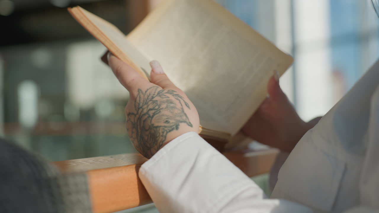 Rear view of fair skinned woman with floral hand tattoo flipping page of novel while reading in natural daylight, relaxed posture by wooden rail, white shirt and blurred modern interior in background