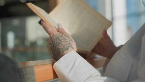 rear view of fair skinned woman with floral hand tattoo flipping page of novel while reading in natural daylight, relaxed posture by wooden rail, white shirt and blurred modern interior in background