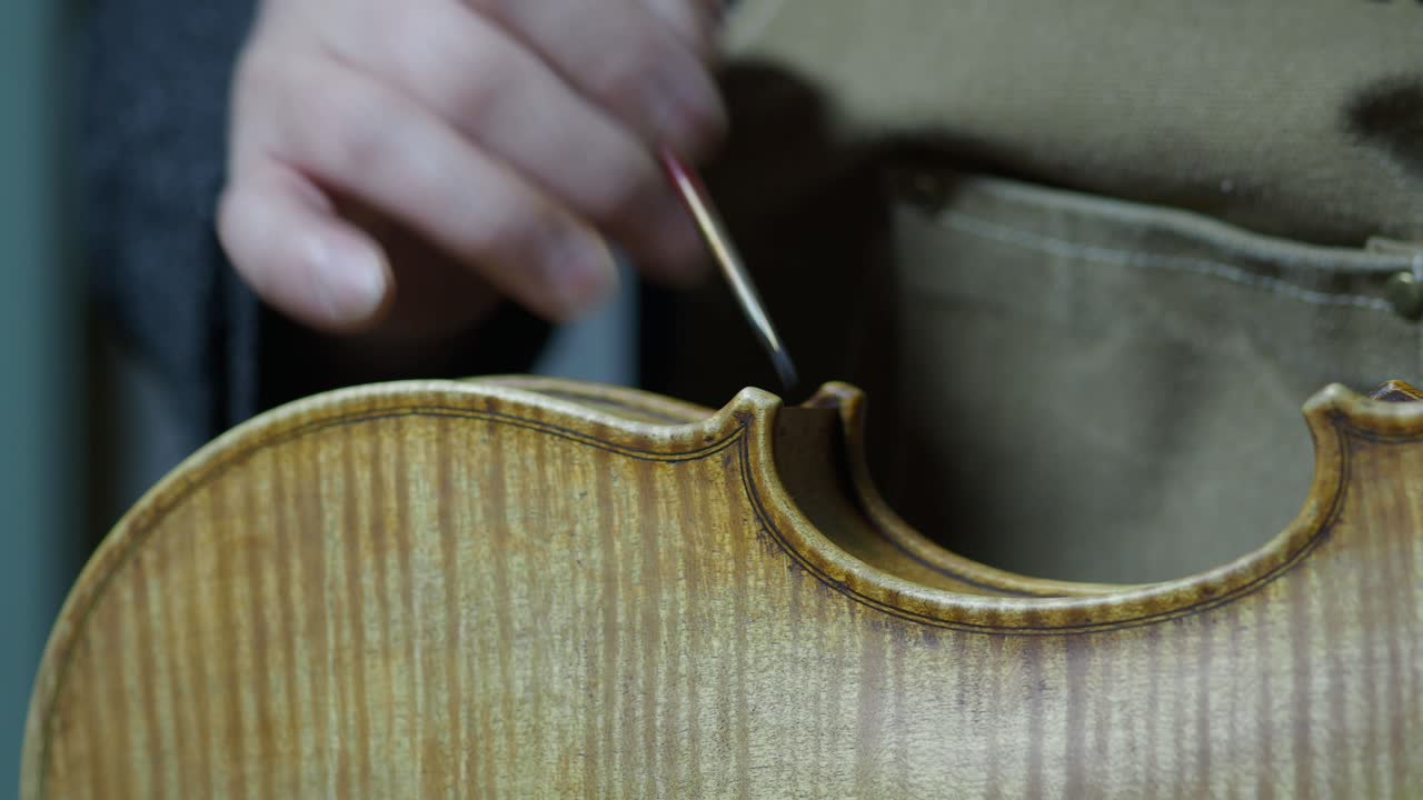 Close-up of a luthier hand-painting varnish onto a violin's ribs, capturing a precise moment in the antiquing process inside a workshop
