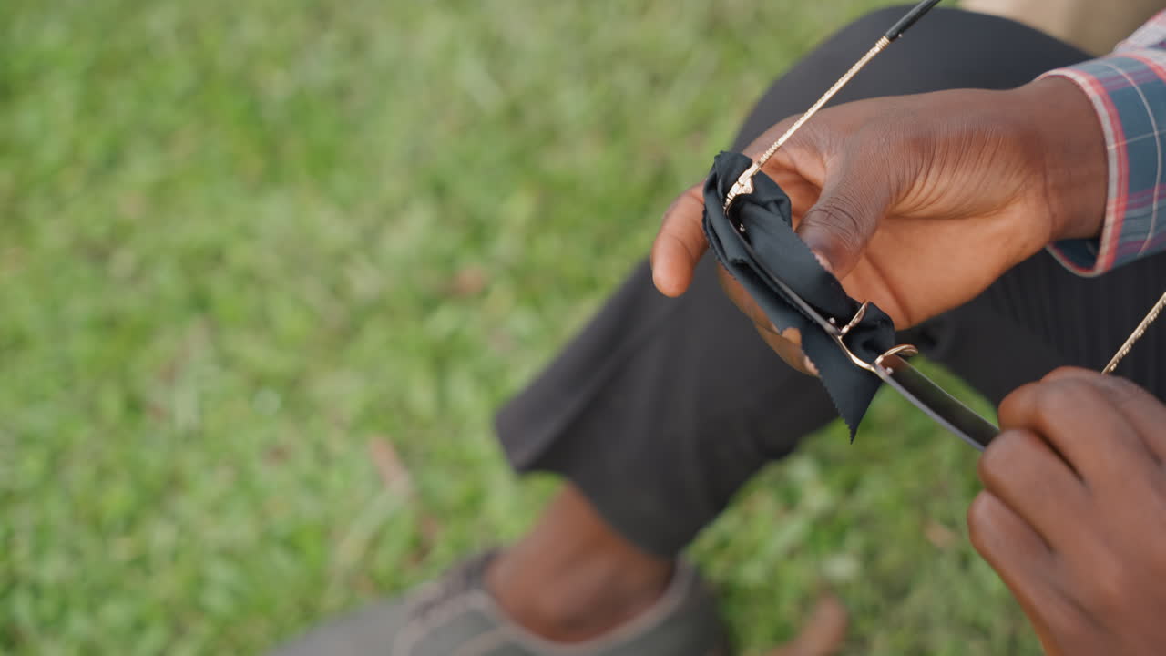 Uomo urbano che lucida le lenti degli occhiali da sole. Uomo rilassato che pulisce gli occhiali da sole riflettenti in un contesto di parco cittadino. Uomo vestito casualmente che ripristina la chiarezza degli occhiali da sole mentre si rilassa su una panchina all'aperto