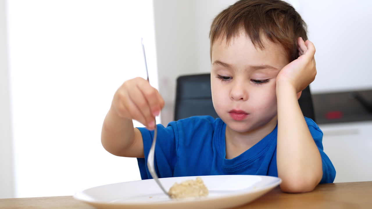 Child is eating a meal at home table. A young boy is sitting at a table, looking thoughtful while eating from a plate with a spoon