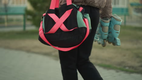 a close-up shot of a girl dressed in a peach jacket and black trousers, walking through a park while carrying a gym bag with bright pink straps and a pair of blue rollerblades