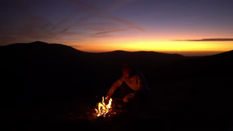 man starts and enjoys bonfire alone at sunset campsite in the nature