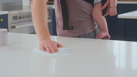 transgender father cleaning kitchen looking after baby son in sling