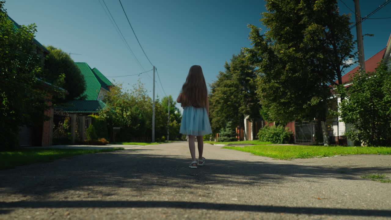 Back view of little girl in white dress with long flowing hair walking down quiet empty suburban street holding teddy bear, surrounded by green trees, houses, and clear blue sky on sunny day
