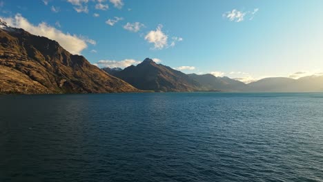 Panorámica-Aérea-De-Camiones-A-Través-Del-Lago-Wakatipu-Se-Extiende-Hasta-Las-Montañas-De-Arenisca-Roja