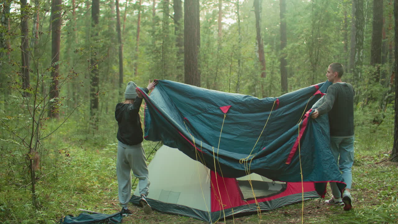 Couple lifts tent cover together to spread over pitched tent during forest camping, surrounded by trees and soft sunlight as they coordinate setup in peaceful natural environment