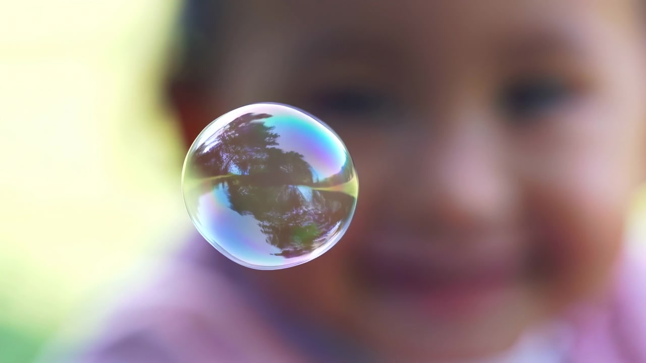 Una burbuja de jabón transparente está centrada en primer plano con un niño pequeño borroso al fondo, capturada con luz natural suave del día con poca profundidad