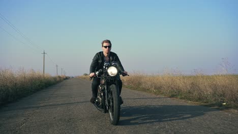 closeup view of a stylish cool young man in sunglasses and leather jacket riding motorcycle on a road on a sunny day. then the camera moves forward and the man visibly shrinking into the distance