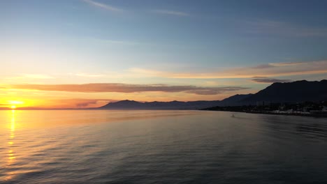 Vista-De-La-Puesta-De-Sol-Del-Mar-De-Marbella-Al-Atardecer,-Filmada-Con-Gimbal-En-Un-Barco-Que-Se-Traslada-Al-Mar-Mediterráneo-Surfista-En-La-Distancia