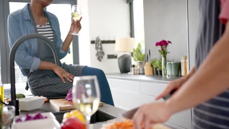 happy diverse couple wearing aprons drinking wine and preparing meal in kitchen, slow motion