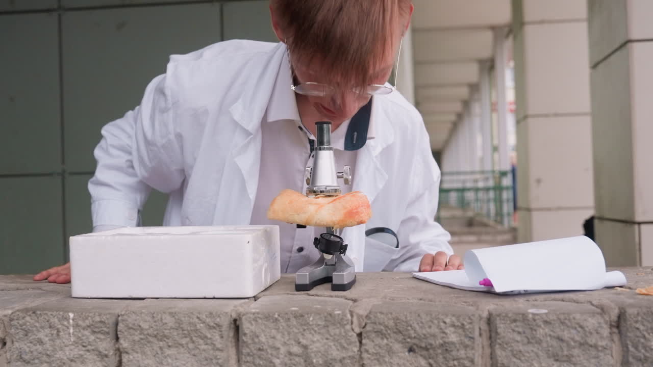 Young biologist in white coat rises up after carefully examining pastry closely through microscope outdoors, reflecting focus, research mindset, and dedication to unusual scientific observation