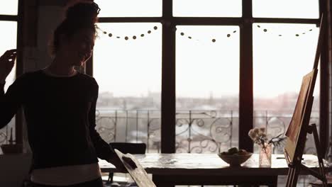 young cute curly female artist dancing in the art studio then finishes up the lines on her painting, with the background of beautiful sunset.