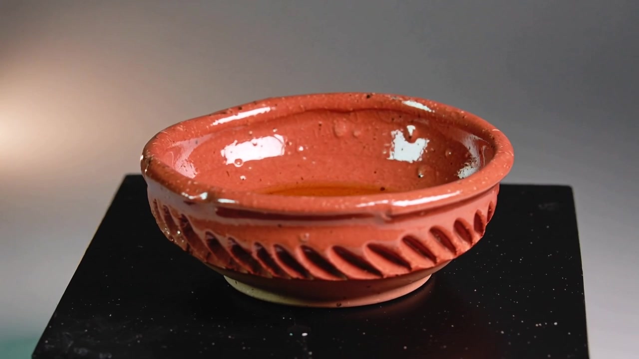 La telecamera si sposta a destra verso una ciotola di terracotta smaltata in studio che mostra un liquido ambrato, una scheggiatura sul bordo e un riflesso.