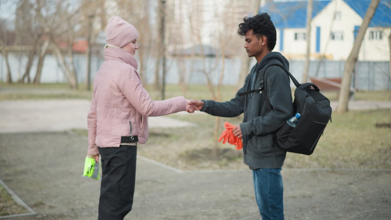 Black guy wearing dark gray hoodie holding orange gloves standing facing woman in pink jacket and beanie holding neon safety vest, meeting outdoors in park during early spring with urban background