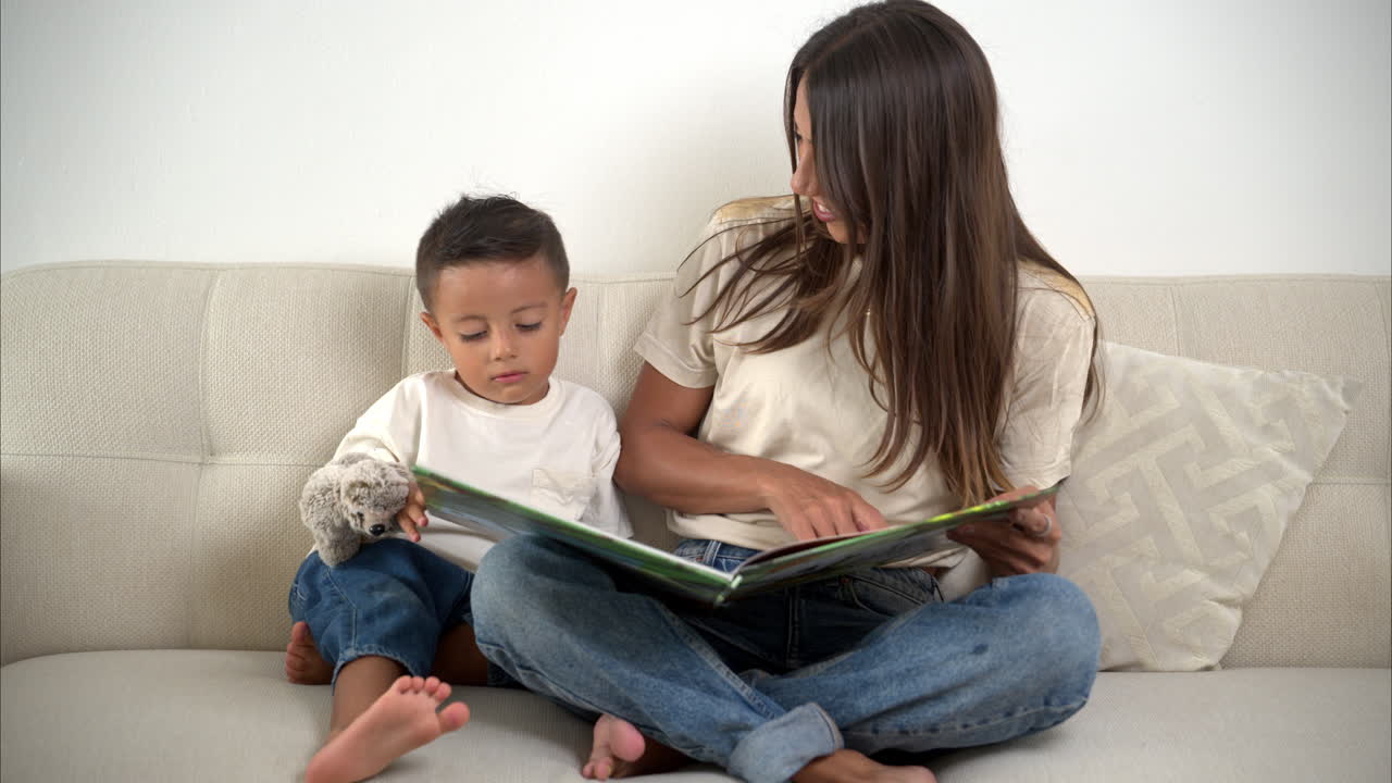 cámara lenta de una madre mexicana latina morena leyendo un libro a su hijo sentada en el sofá con una camiseta beige y pantalones vaqueros azules