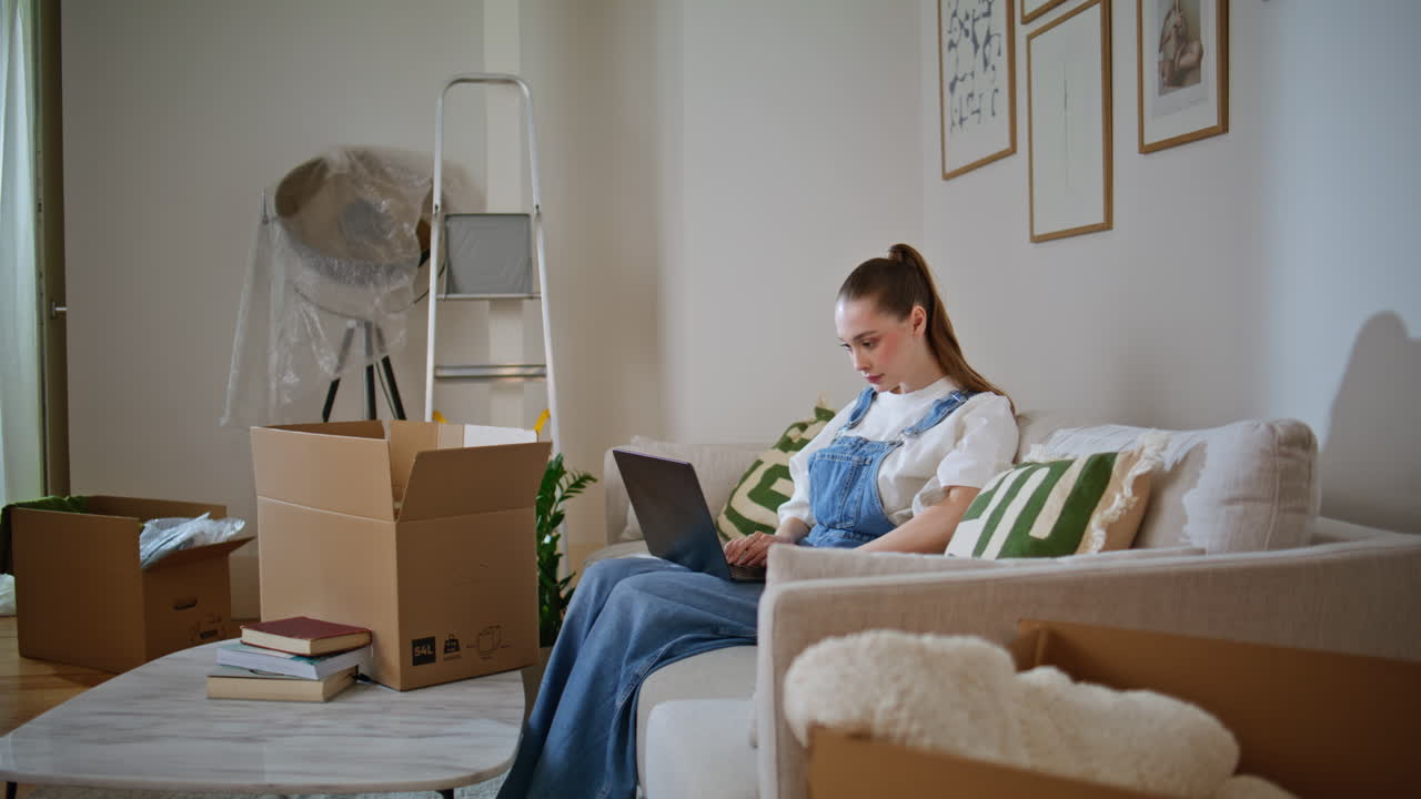 New tenant looking laptop sitting couch near moving cardboard boxes. Woman home