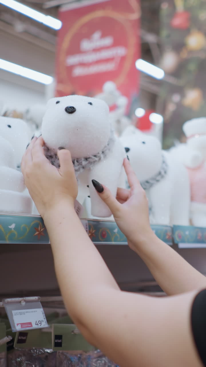 검은 셔츠와 안경을 입은 긴 갈색 머리카락을 가진 젊은 여성의 측면 뷰, 색 플러시 북극  장난감과 스카프가 가게 선반에 축제 휴가 장식으로 둘러싸여 있습니다.