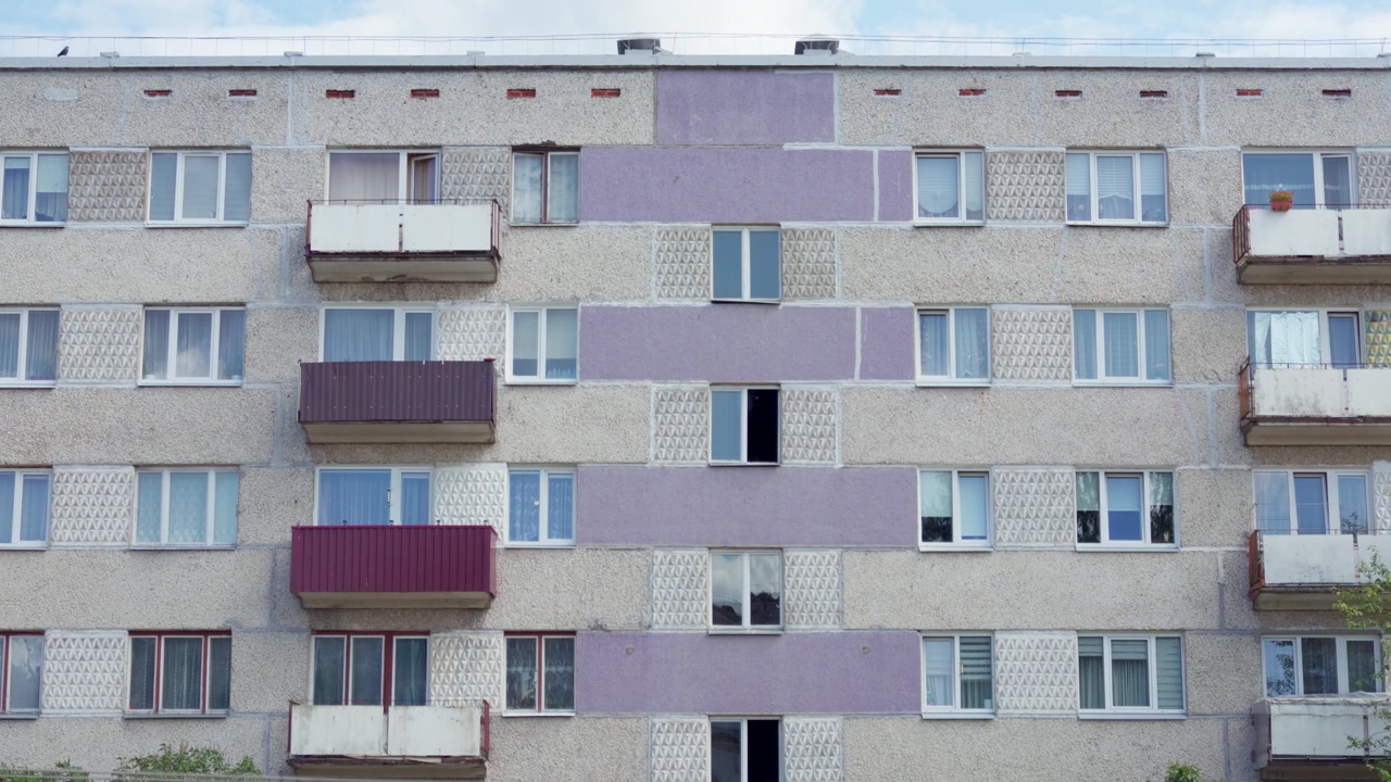 Statický střední záběr ukazující fasádu panelového domu ze sovětské éry s balkony v Daugavpilsu za jasného letního dne, odrážející kontext městského baltského bydlení.