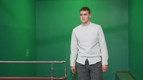 front shot of male in white shirt and checkered trousers walking up stairs, holding rail. focused expression under green lighting. stylish indoor setting with modern metal railing and green walls