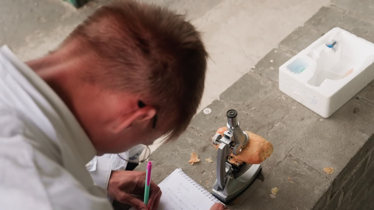 Back view of young man in white coat carefully examining pastry through microscope in outdoor setting, reflecting concentration, curiosity, and scientific observation during dedicated research study