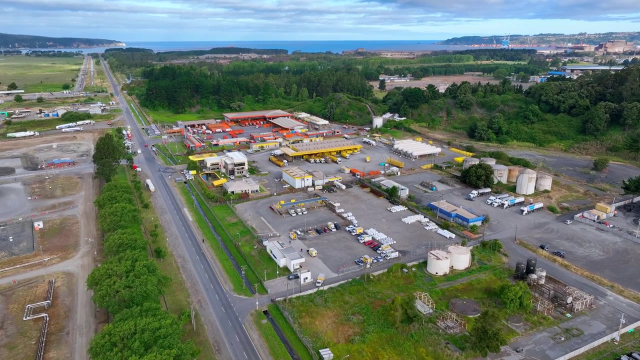 Vuelo hacia adelante sobre un complejo logístico industrial con almacenes coloridos, tanques de almacenamiento y una autopista, revelando la costa del Océano Pacífico y las instalaciones portuarias a lo lejos. Hualpén, Biobío, Chile.