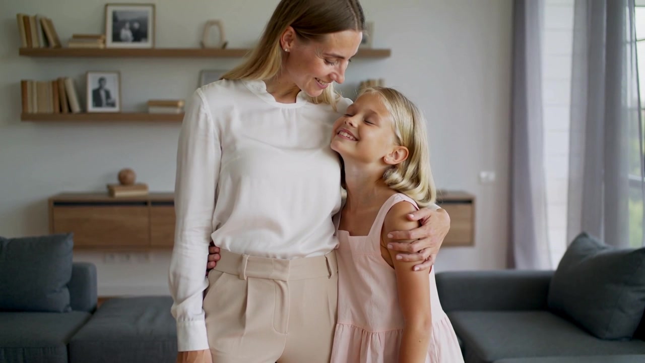 Mutter in weißer Bluse dreht den Kopf und tröstet Kind in rosa Kleid im Wohnzimmer, graues Sofa.