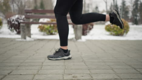 close-up leg view of athlete jogging in black sneakers and leggings along snowy park pathway, with shoelace swaying during motion, surrounded by frosty winter scenery, benches, and colorful bushes