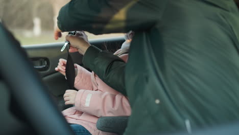 a man in a green jacket is securing the seat belt for his young daughter, who is wearing a pink jacket and a beanie, inside a car. the child looks attentively at her father while he ensures her safety