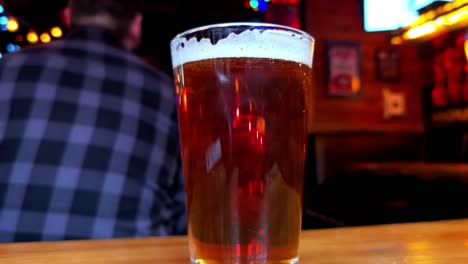 hipster-seated-pub-beer-closeup-bokeh-background-foreground-with-wooden-wall-siding-TV-screen-closeup-of-lager-draft-with-foam-and-clear-table-inside-public-gathering-of-great-atmosphere-vintage-art