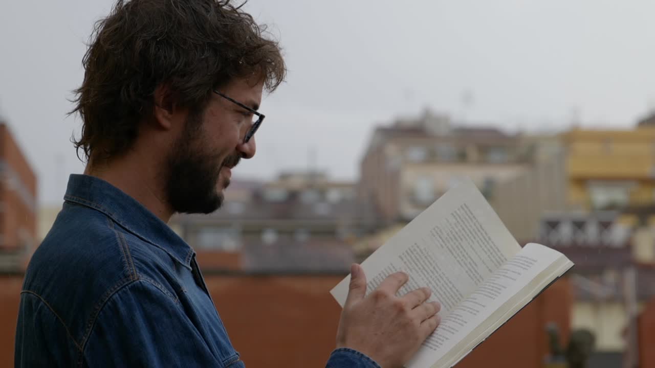 vista lateral de un hombre barbudo feliz leyendo un libro bajo la lluvia