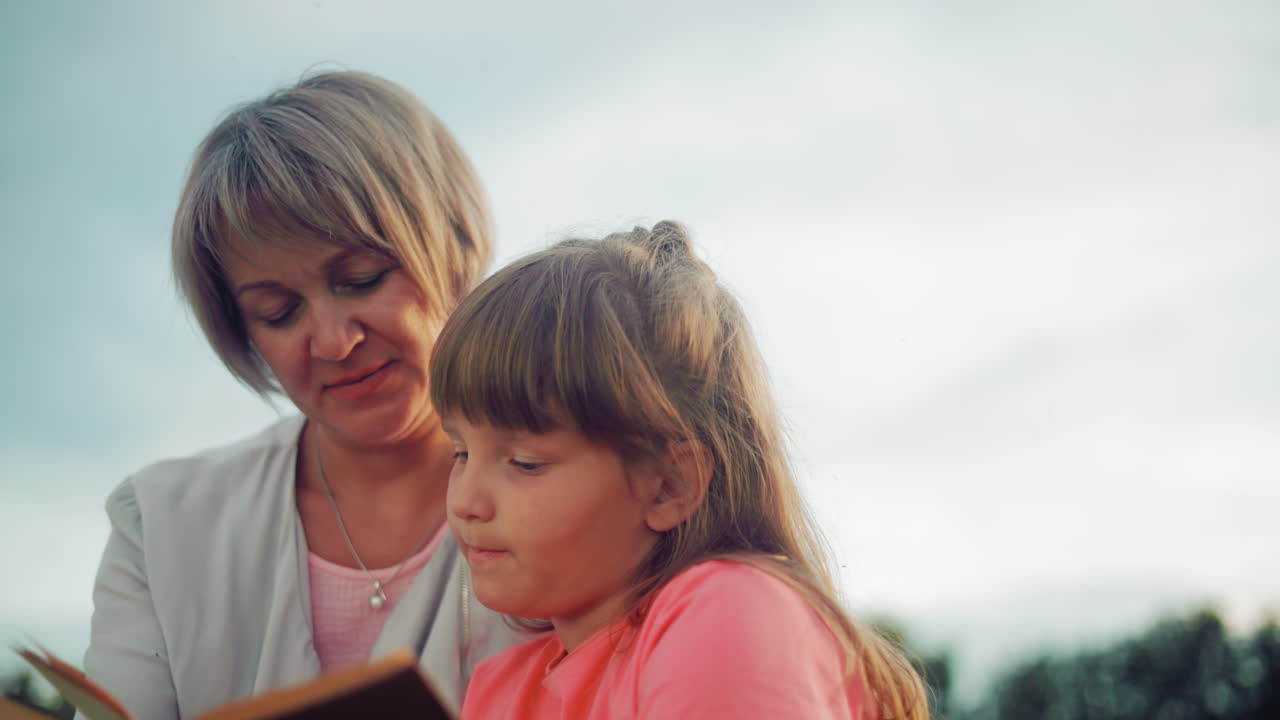 Eine zärtliche Szene einer Mutter in einer weißen Jacke, die ihre Tochter in Rosa umarmt, während sie gemeinsam im Freien lesen und einen Moment der Bildung und Wärme teilen.