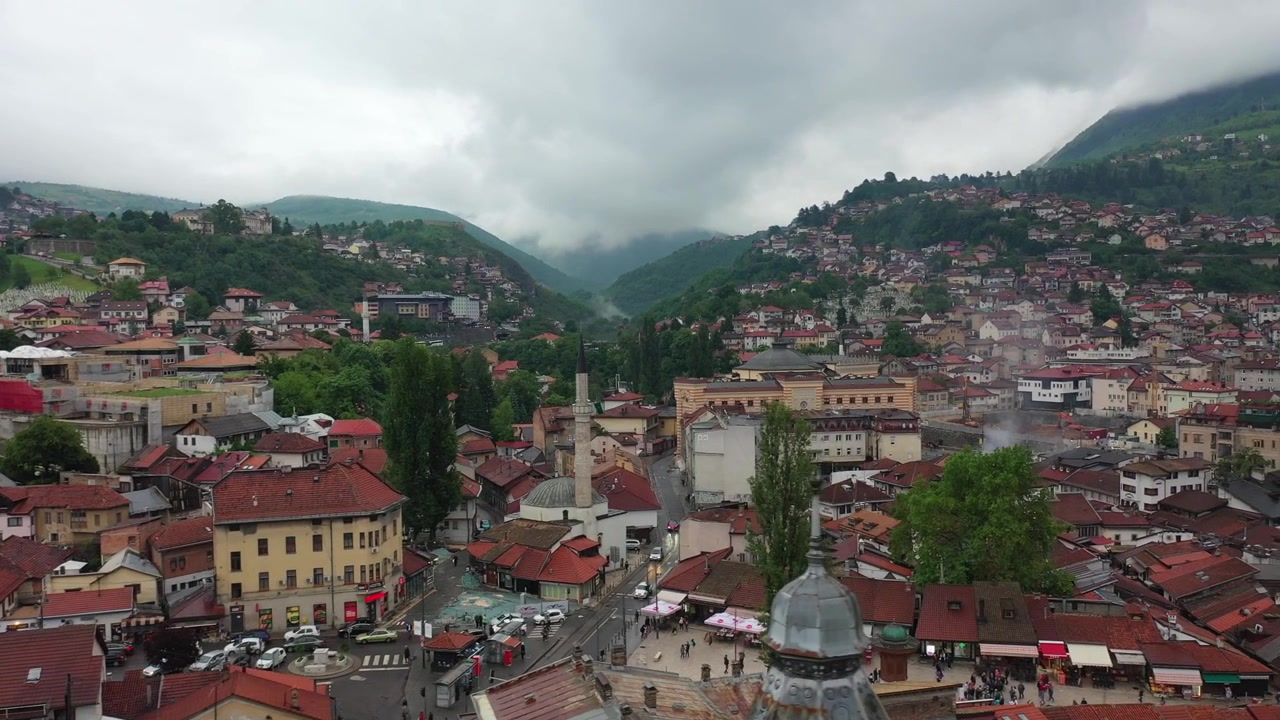Cảnh quay drone trên cao góc rộng cho thấy sự mở rộng đô thị của Sarajevo, nổi bật với những mái nhà đỏ, các tòa nhà cao tầng hiện đại và những đám mây thấp bao phủ các ngọn núi xa ở Bosnia và Herzegovina