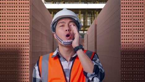 close up of asian male engineer with safety helmet yelling with hand over mouth while standing with red brick packed in stacks are stored