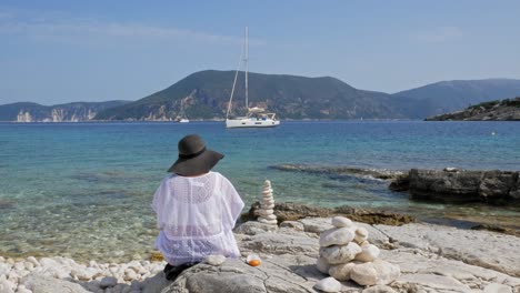 back view of a girl sitting on the rocky coast of emplisi beach with yachts anchored in the ocean in the background