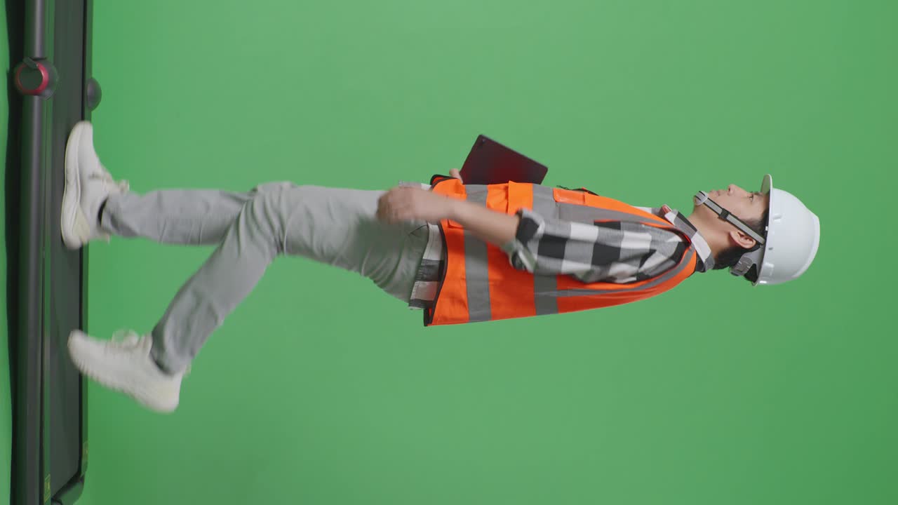 vista lateral de cuerpo completo ingeniero masculino asiático con casco de seguridad sosteniendo una tableta mientras camina en el estudio de fondo de pantalla verde